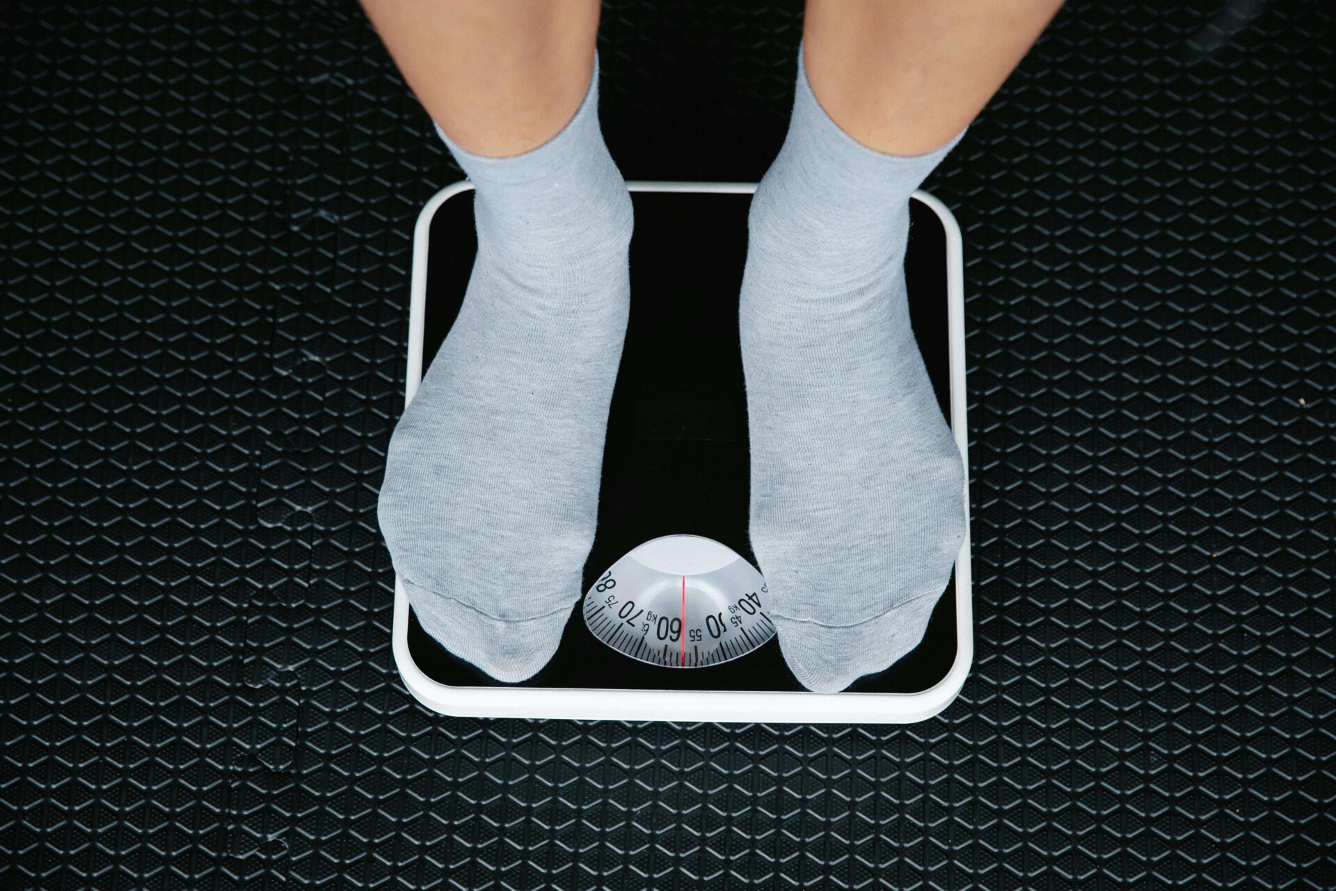 Person wearing gray socks standing on a black analog weighing scale with a white border, placed on a textured black floor. The scale's dial is visible, showing the weight measurement.